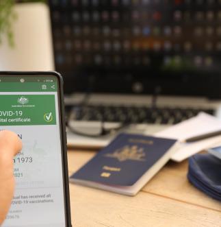 A person sits at their desk in front of their laptop and Australian passport, holding a phone which shows their COVID-19 digital certificate.